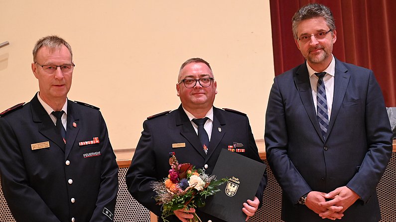 Auf dem Bild sind drei Personen zusehen. Die Person in der Mitte hält eine Urkunde und einen Blumenstrauß in den Händen. 