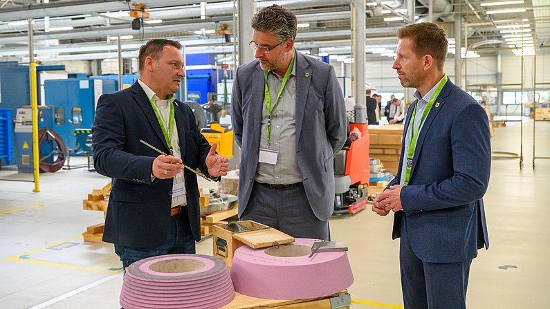 Drei Männer stehen in einer Fabrik und schauen sich an. Einer hält ein Sägeblatt in der Hand. Im Vordergrund sind runde Schleifsteine zu sehen.