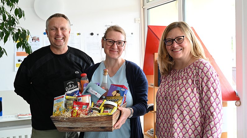 Auf dem Foto sind drei Personen zu sehen. Eine Frau hält einen Präsentkorb vor sich und lächelt