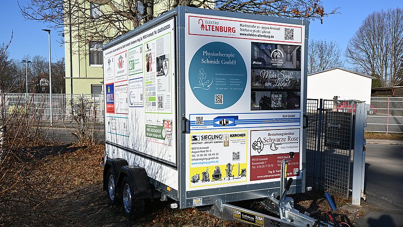 Auf dem Foto ist eine Fahrzeuganhäger zu sehen. Auf der Plane des Anhängers sind viele Firmenlogos zu sehen. 