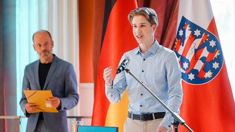 Auf dem Bild sind zwei Personen zu sehen. Die eine Person spricht am Mikrofon, die andere steht hinter ihm. 