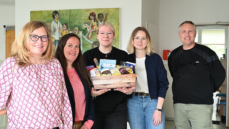 Auf dem Foto sind fünf erwachsene Personen zu sehen. Eine Frau hält einen Präsentkorb vor sich. Alle lächeln. 