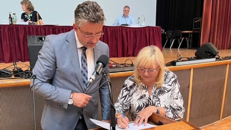 Ein Mann und eine Frau stehen am Pult. Sie unterschreibt einen Zettel.