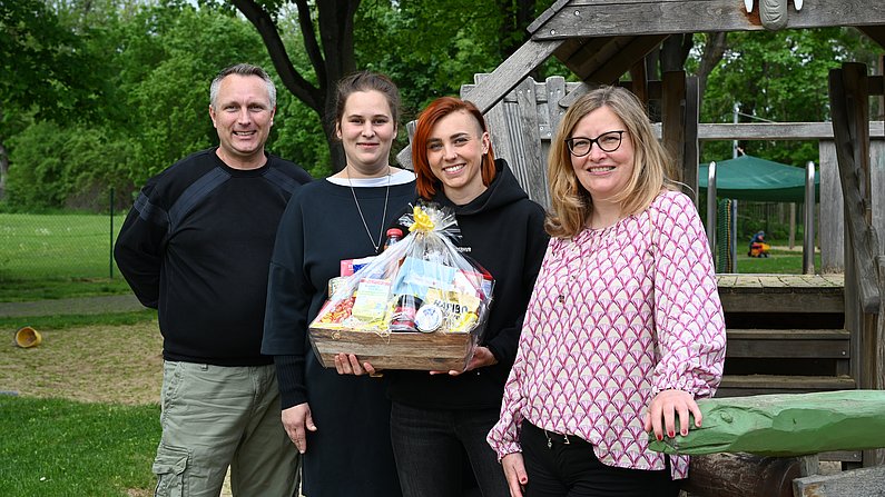 Auf dem Foto sind vier Erwachsene zu sehen. Eine Frau hält einen Präsentkorb vor sich. Alle lächeln. 