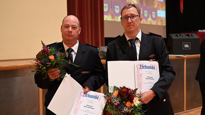 Auf dem Bild sind zwei Personen zu sehen. Beide halten eine Urkunde und einen Blumenstrauß in den Händen. 